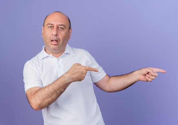 Impressed casual mature businessman looking at camera pointing at side isolated on purple background