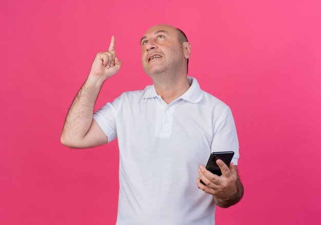 Impressed casual mature businessman looking and pointing up and holding mobile phone isolated on pink background with copy space