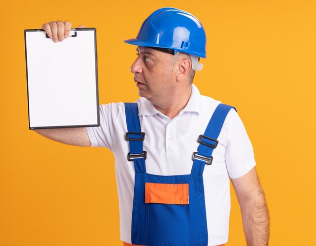 Impressed caucasian adult builder man in uniform holds and looks at clipboard on orange