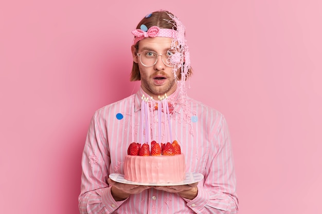 Uomo caucasico impressionato reagisce a qualcosa di sorprendente festeggia il compleanno andando a soffiare candele ed esprimere il desiderio vestito con abiti eleganti isolati sopra il muro rosa