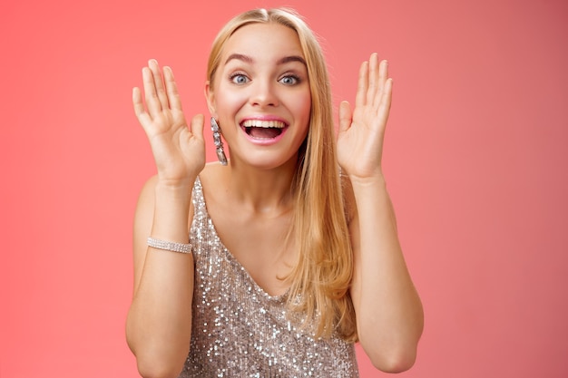 Impressed charming entertained blond european 25s woman in stylish silver glamour dress having fun playing peekaboo smiling joyfully hold palms near face laugh amused, standing red background