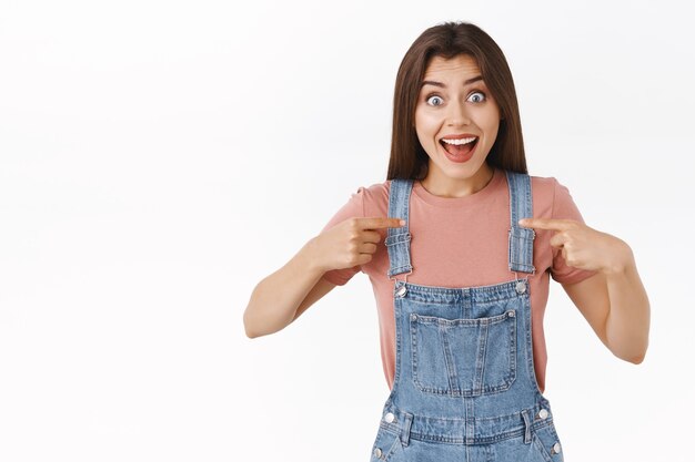 Impressed, cheerful enthusistic brunette woman in denim overalls, t-shirt, pointing herself with astonished surprised expression, smiling broadly, was chosen, winning, receive prize, white background