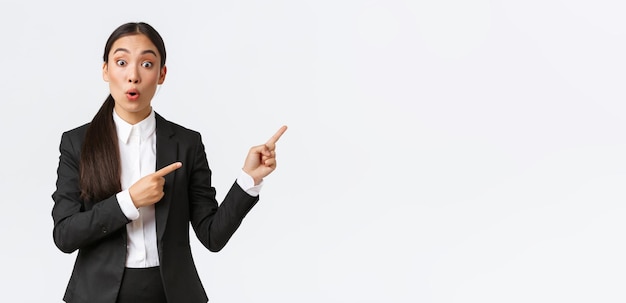 Free photo impressed and curious asian female entrepreneur asking question about project businesswoman in black suit pointing fingers right to get all details about product look astounded white background