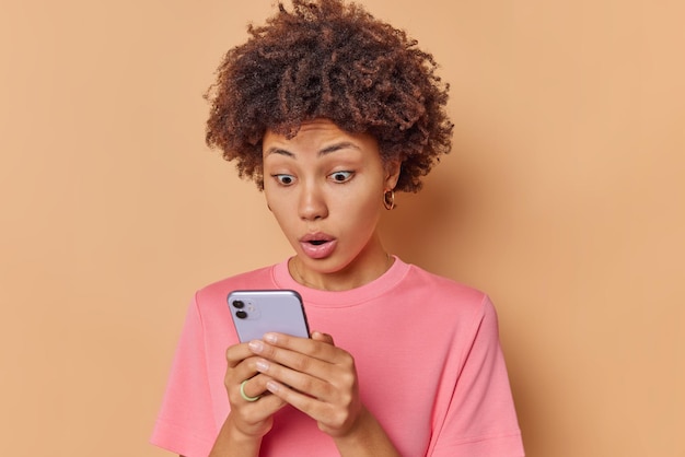 Впечатленная кудрявая женщина получает шокирующее сообщение, смотрит на смартфон, широко открывает рот, одетая в повседневную футболку, изолированную на бежевом фоне, обнаруживает удивительные новостные киоски в помещении