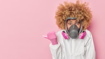 Впечатленная кудрявая женщина-ученый носит костюм для защиты от коронавируса, а маска указывает большим пальцем на макет пространства, изолированного на розовом фоне, демонстрирует рекламу защита от биологической опасности