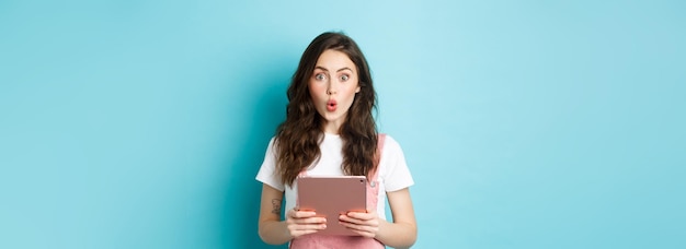 Free photo impressed cute girl say wow stare excited at camera holding digital tablet over chest standing in spring clothes against blue background