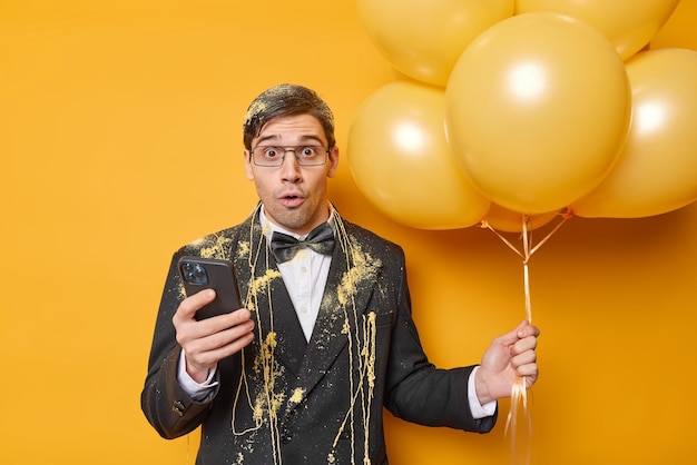 Впечатленный темноволосый мужчина носит праздничную одежду, держит мобильный телефон, а связка гелиевых шаров празднует особый случай, вечеринка изолирована на желтом фоне Концепция праздников людей