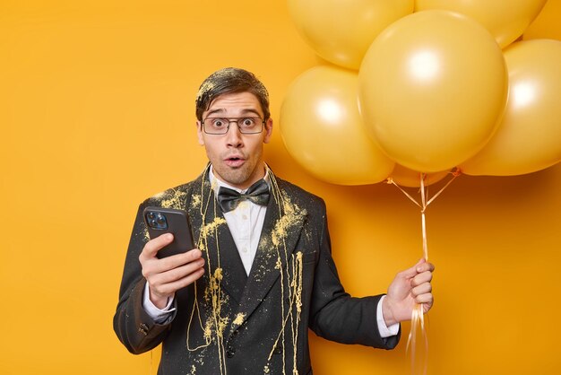 Impressed dark haired man wears festive clothes holds mobile phone and bunch of helium balloons celebrates special occasion has party isolated over yellow background People holidays concept