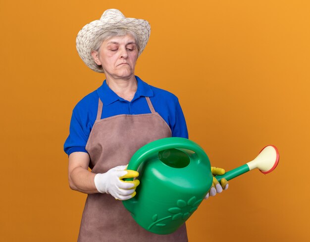 Impressed elderly female gardener wearing gardening hat and gloves holding and looking at watering can isolated on orange wall with copy space