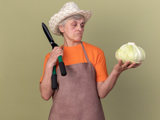 Impressed elderly female gardener wearing gardening hat holding gardening scissors and looking at cabbage on olive green