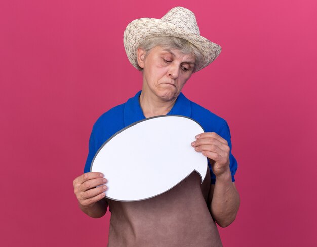 Impressed elderly female gardener wearing gardening hat holding and looking at speech bubble