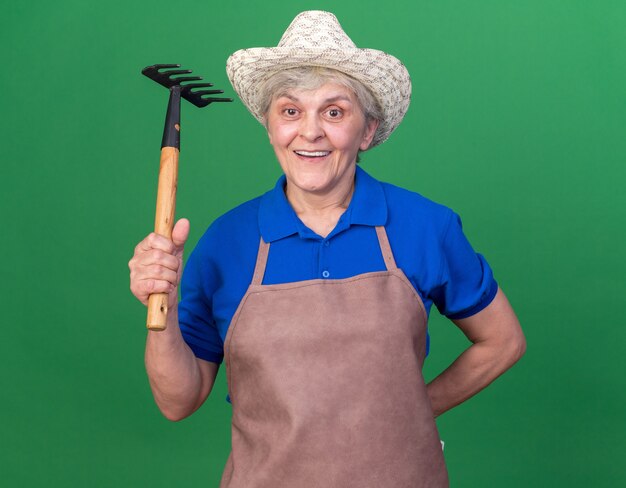 Impressed elderly female gardener wearing gardening hat holding rake isolated on green wall with copy space