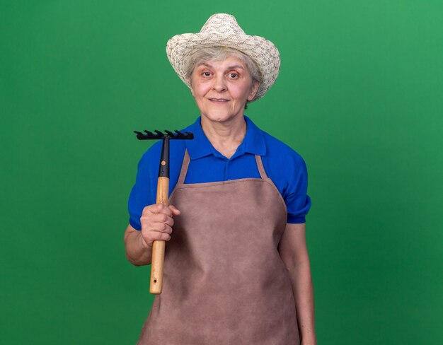 Impressed elderly female gardener wearing gardening hat holds rake