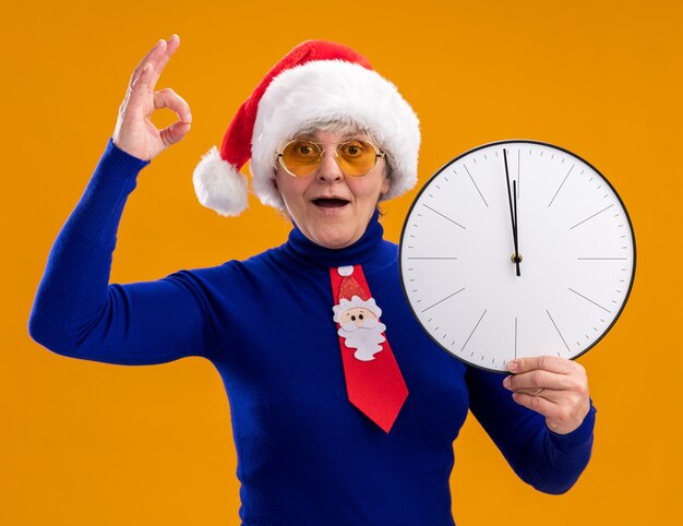 Foto gratuita la donna anziana impressionata in occhiali da sole con cappello da babbo natale e cravatta da babbo natale tiene l'orologio e gesticolando segno ok isolato sulla parete arancione con spazio di copia