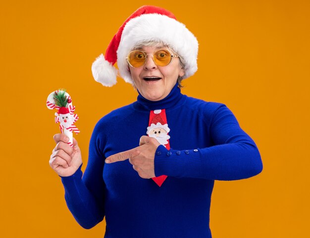 Впечатленная пожилая женщина в солнцезащитных очках в шляпе санта-клауса и галстуке санта-клауса держит и указывает на конфету, изолированную на оранжевом фоне с копией пространства