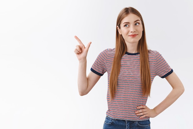 Impressed, enthusiastic good-looking caucasian woman in striped t-shirt, tilt head and smirk amused, pointing looking upper left corner, check out something interesting and cool, white background