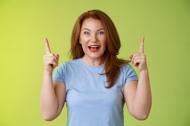 Impressed enthusiastic happy cheerful middleaged mature woman red hair smiling astonished look admiration joy pointing up raised index fingers introduce impressive stunning offer green wall