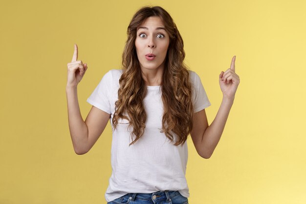 Impressed enthusiastic surprised ambushed cute female model fold lips wow admiration joy stare camera fascinated pointing up index fingers top copy space show you stunning offer yellow background