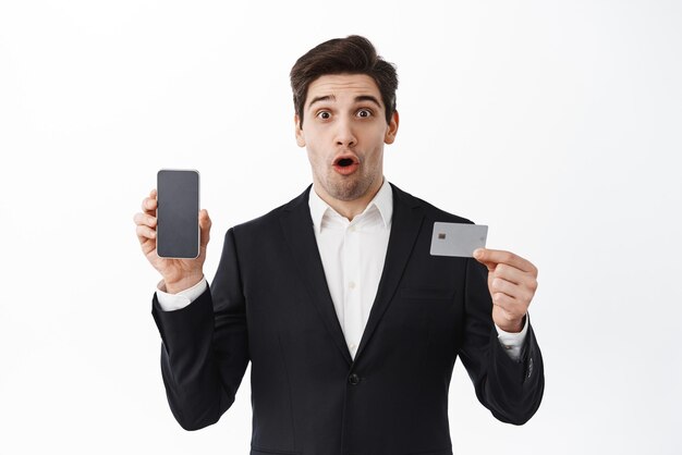 Impressed entrepreneur showing phone and credit card gasping amazed standing over white background in black suit