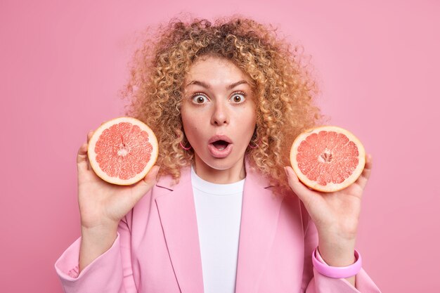 Впечатленная европейская женщина держит две половинки свежего грейпфрута, удивленная, узнав о полезных веществах, которые содержит этот цитрусовый фрукт, и вьющиеся волосы изолированы на розовой стене. На диете