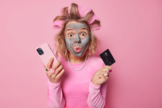Впечатленная европейская женщина с маской красоты на лице применяет бигуди для волос, делает прическу, держит мобильный телефон и кредитную карту для покупок в Интернете, готовится к свиданию на розовом фоне