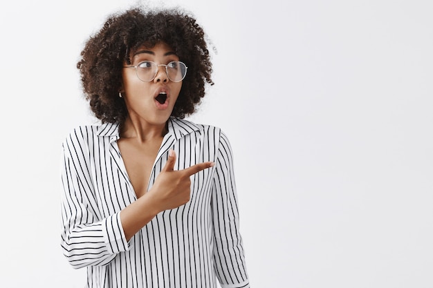 impressed and excited african american businesswoman with afro hairstyle in glasses and striped blouse gasping while looking and pointing right with amazement and interest