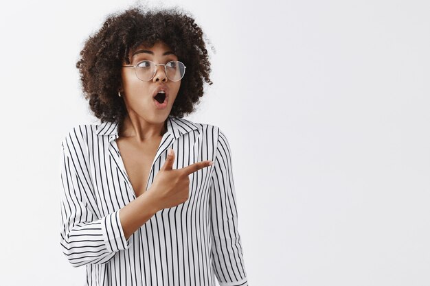 impressed and excited african american businesswoman with afro hairstyle in glasses and striped blouse gasping while looking and pointing right with amazement and interest