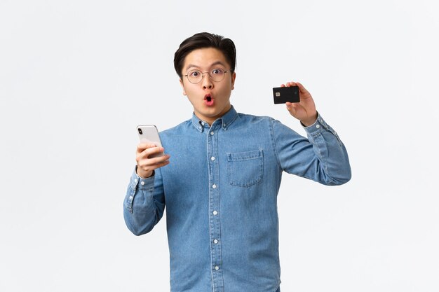 감동적이고 흥분한 아시아 은행 고객, 안경을 쓰고 신용카드를 보여주는 캐주얼 옷을 입은 남자, 스마트폰을 사용하여 전자 뱅킹 응용 프로그램을 열고, 모바일 앱으로 인터넷에서 쇼핑합니다.