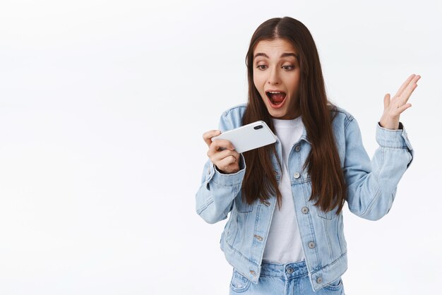 スマートフォンで叫んでいる感動興奮とスリル満点の若い女の子は、携帯電話を水平に見つめている画面を驚かせた白い背景技術の概念を持って手を上げて勝利を上げて驚いた