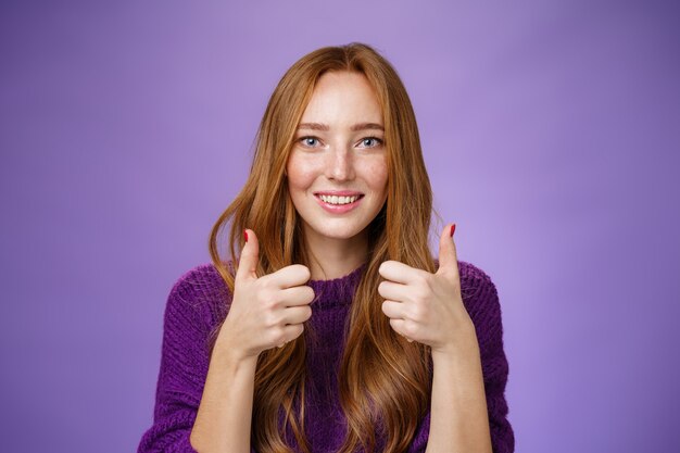 Impressed girl admiring great music taste of friend showing thumbs up and smiling excited and astonished expressing like and positive feedback, approving and supporting plan over purple background.