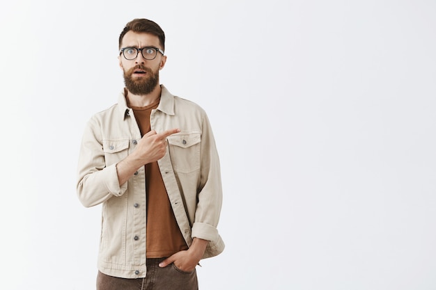 Impressed handsome bearded man in glasses posing against the white wall