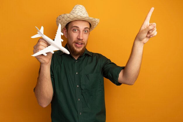 Impressed handsome blonde man with beach hat holds model plane and points up isolated on orange wall