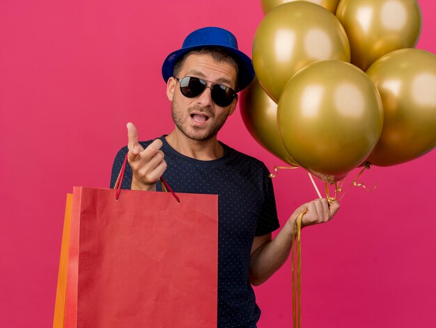 Impressed handsome caucasian man in sun glasses wearing blue party hat holds helium balloons and paper shopping bags pointing at camera isolated on pink background with copy space