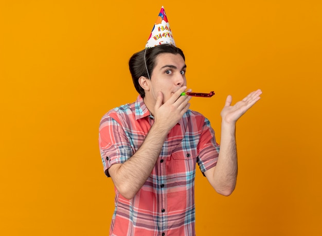 Impressed handsome caucasian man wearing birthday cap holding hand open and blowing party whistle 