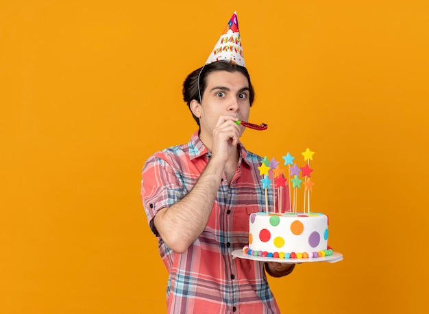 Impressed handsome caucasian man wearing birthday cap holds birthday cake blowing party whistle