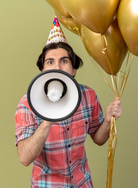 Impressed handsome caucasian man wearing birthday cap holds helium balloons and speaks into loud speaker 