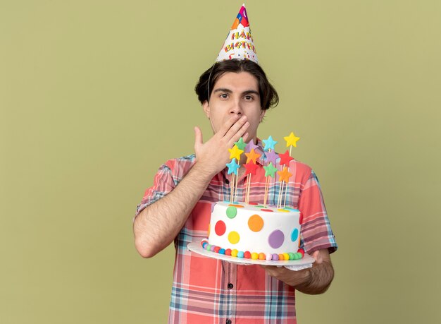 Impressed handsome caucasian man wearing birthday cap puts hand on mouth and holds birthday cake 