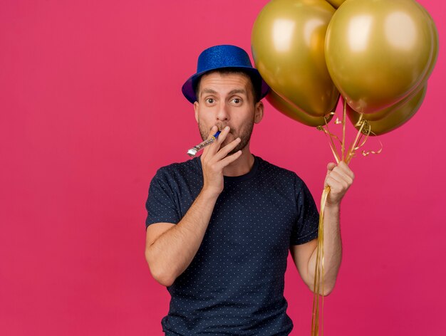 Impressed handsome caucasian man wearing blue party hat holds helium balloons blowing whistle isolated on pink background with copy space