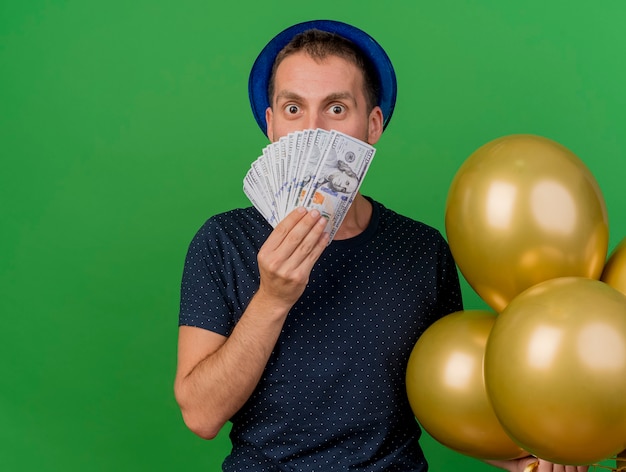 L'uomo caucasico bello impressionato che porta il cappello blu del partito tiene i palloni dell'elio e soldi isolati su fondo verde con lo spazio della copia