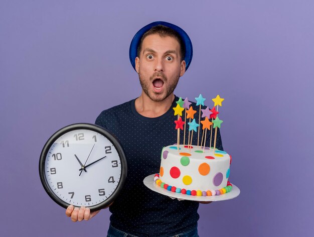 Impressed handsome man wearing blue hat holds birthday cake and clock isolated on purple wall with copy space