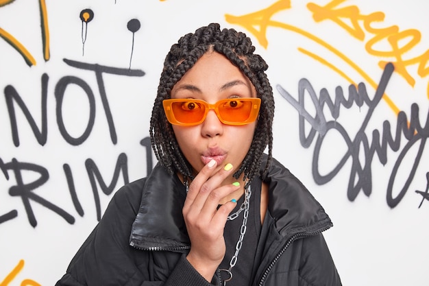 Impressed hipster teenage girl with dreadlocks looks speechless at camera keeps hand on folded lips wears trendy orange sunglasses and black jacket poses against graffiti wall