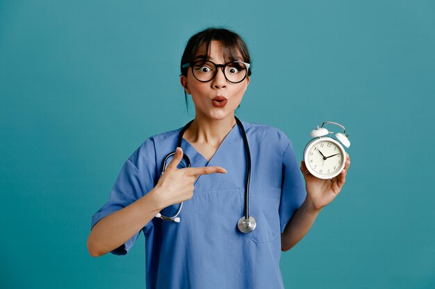 Впечатлен, держа будильник молодая женщина-врач в униформе фит стетоскоп, изолированные на синем фоне