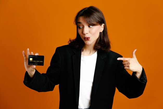 オレンジ色の背景に分離された黒いジャケットを着ている若い美しい女性に感銘を受けた保持カード