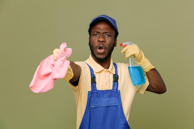 Впечатленный держащий чистящее средство с тряпкой молодой афроамериканец-уборщик мужчина в униформе с перчатками, изолированными на зеленом фоне