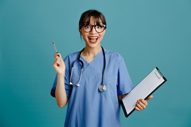 Впечатлен держа буфер обмена с ручкой молодая женщина-врач в форме стетоскоп, изолированные на синем фоне