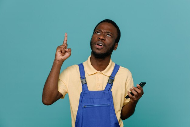 Впечатленный держащий телефон молодой афроамериканец-уборщик в униформе, изолированный на синем фоне