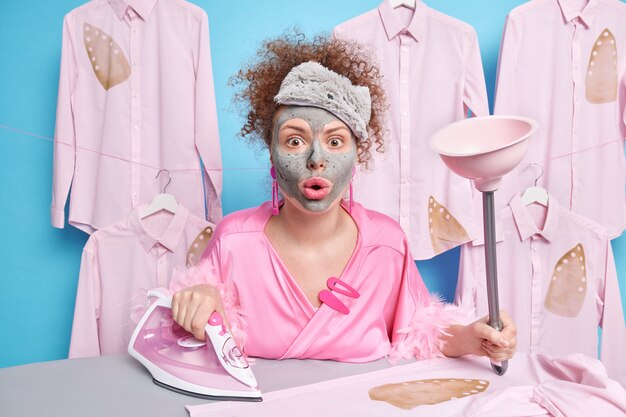 Impressed housewife stares surprised cannot believe in shocking news uses electric iron and plunger for cleaning toilet applies clay mask on face poses against ironing board. Household duty concept