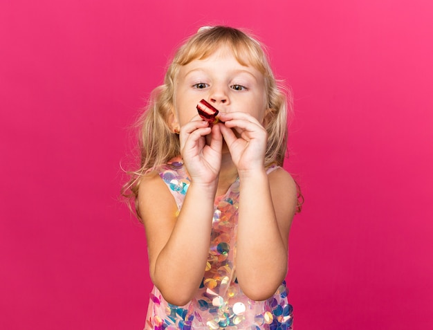 impressed little blonde girl blowing and looking at party whistle isolated on pink wall with copy space