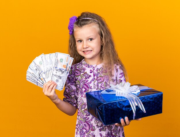 impressed little blonde girl holding gift box and money isolated on orange wall with copy space