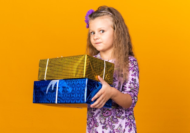 Free photo impressed little blonde girl holding gift boxes isolated on orange wall with copy space